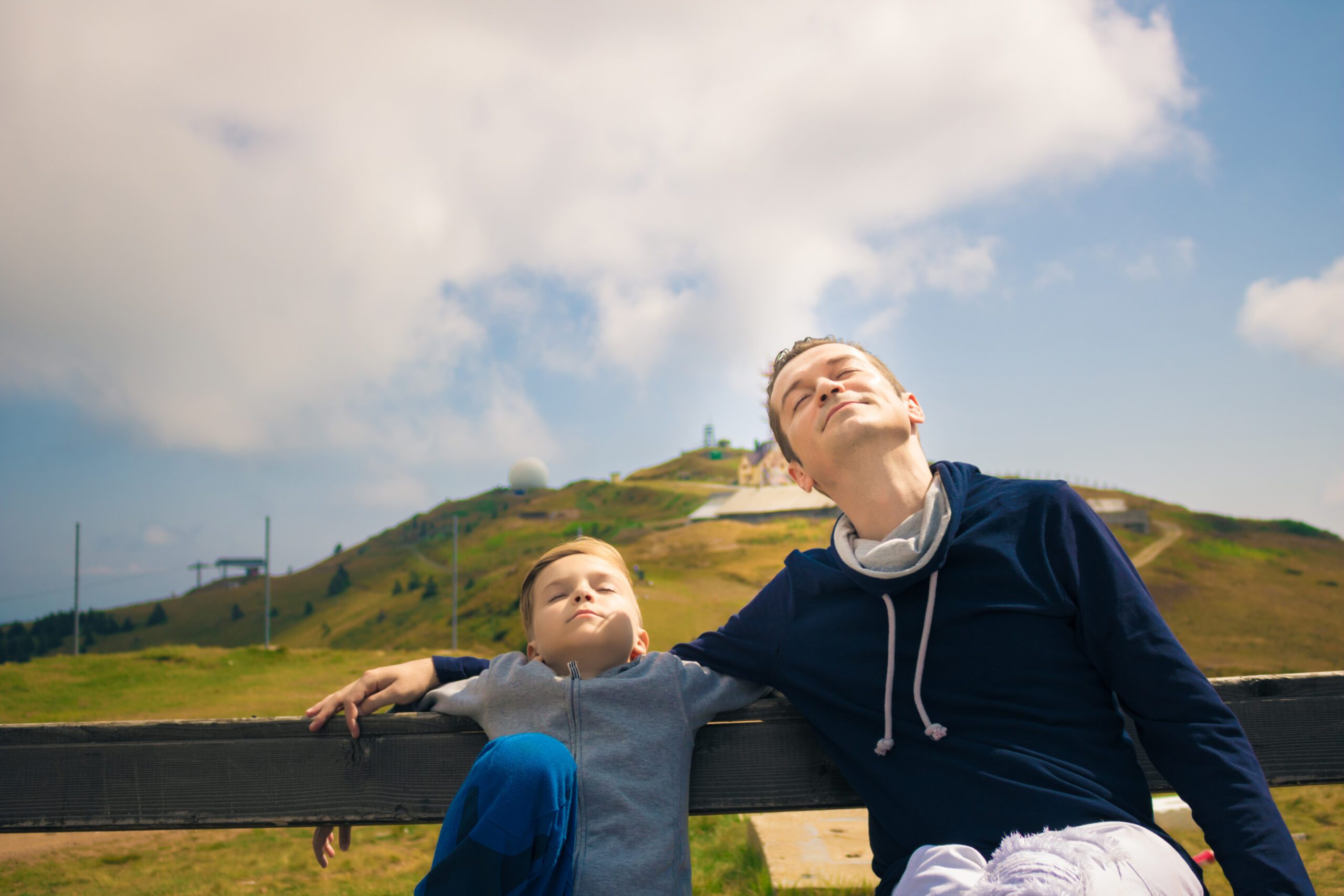 Relaxed father and son enjoying in sunshine while sitting on a bench with eyes closed.