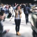 Woman standing still in a busy crowd with her hands covering her face, while the background is motion-blurred to convey a sense of panic or anxiety.