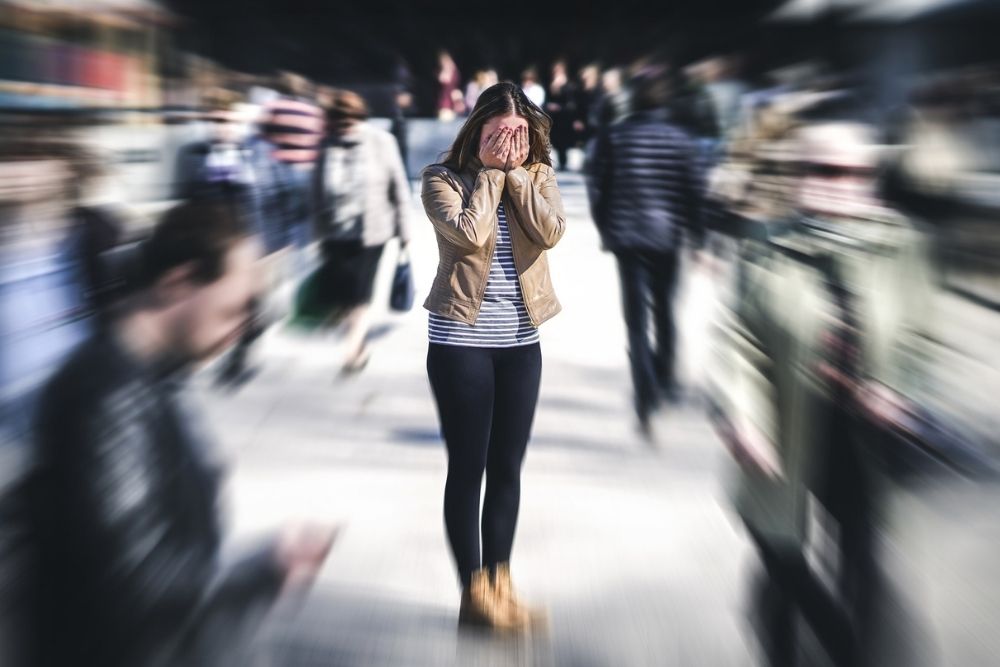 Woman standing still in a busy crowd with her hands covering her face, while the background is motion-blurred to convey a sense of panic or anxiety.
