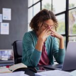 Stressed woman sitting at a desk with her eyes closed and hands on her face, surrounded by papers and a laptop, showing signs of burnout at work.