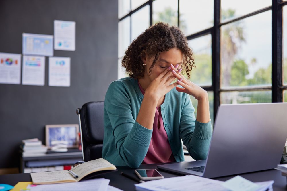 Stressed woman sitting at a desk with her eyes closed and hands on her face, surrounded by papers and a laptop, showing signs of burnout at work.