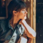 Woman sitting indoors with her chin resting on her hand, gazing thoughtfully out a window with sunlight illuminating her face.