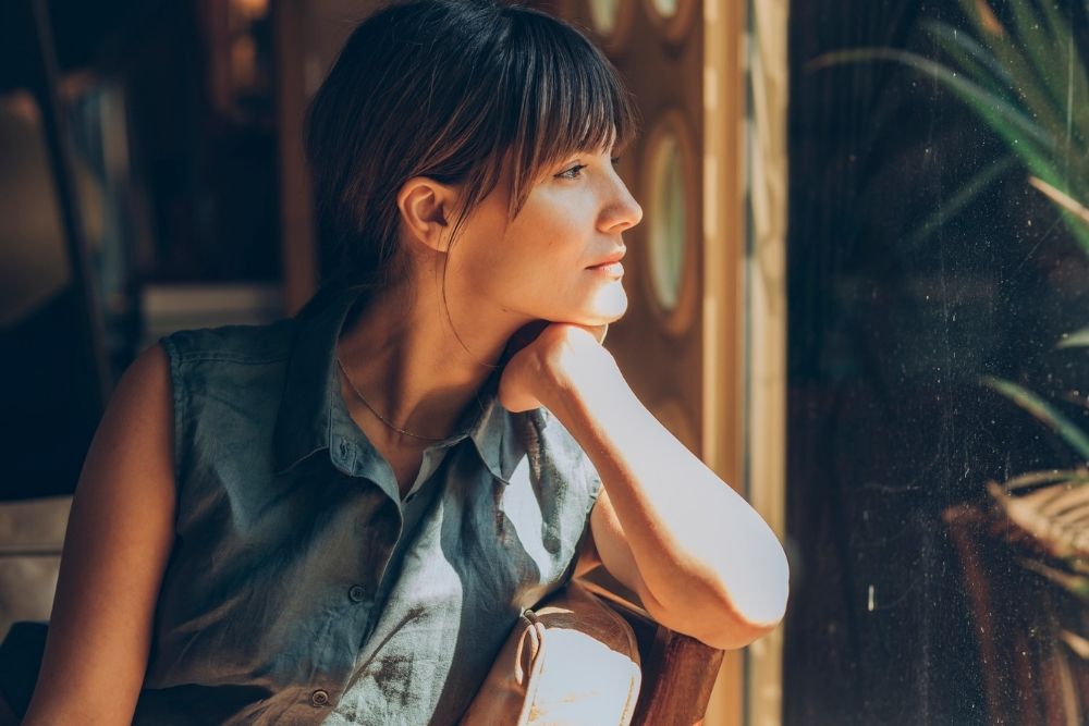 Woman sitting indoors with her chin resting on her hand, gazing thoughtfully out a window with sunlight illuminating her face.