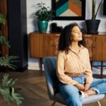A young woman with curly hair sits cross-legged in a chair with her eyes closed, appearing calm and relaxed. She is in a cozy room with houseplants and wooden furniture, practicing mindfulness or meditation.