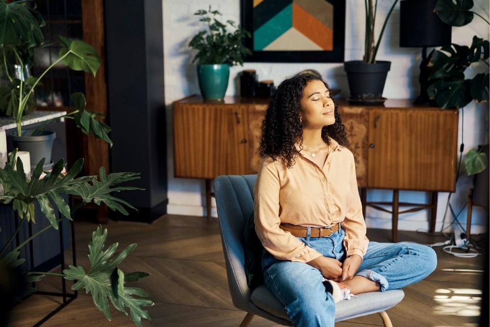 A young woman with curly hair sits cross-legged in a chair with her eyes closed, appearing calm and relaxed. She is in a cozy room with houseplants and wooden furniture, practicing mindfulness or meditation.