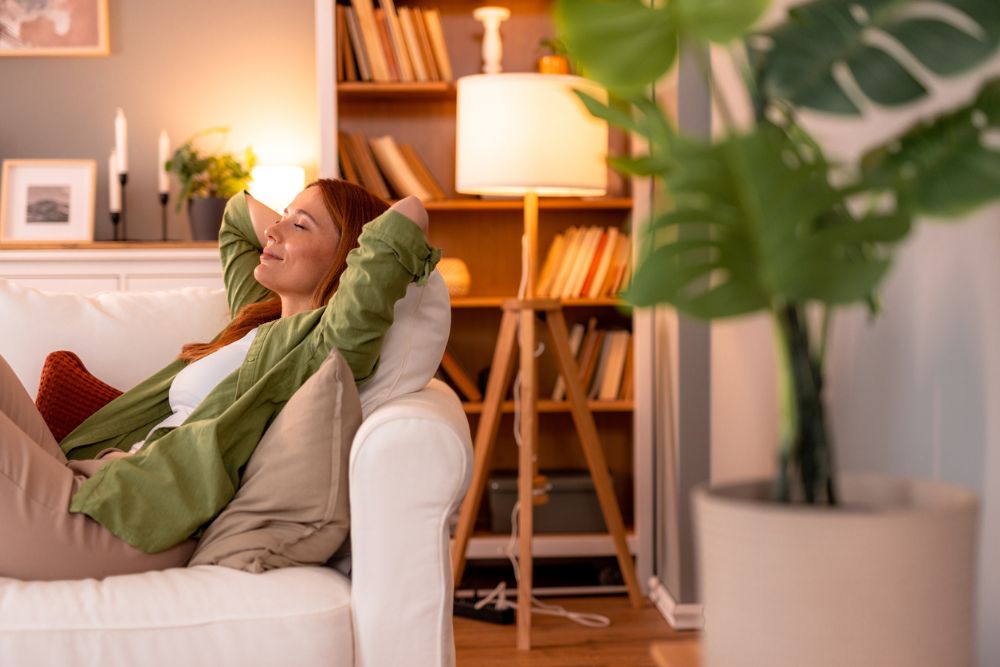 Woman relaxing on a white couch at home, eyes closed and hands behind her head, enjoying a peaceful moment of rest.