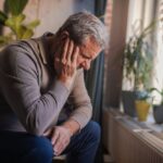 An older man sits indoors with his head resting on his hand, looking down with a distressed expression, symbolizing depression and emotional struggle.