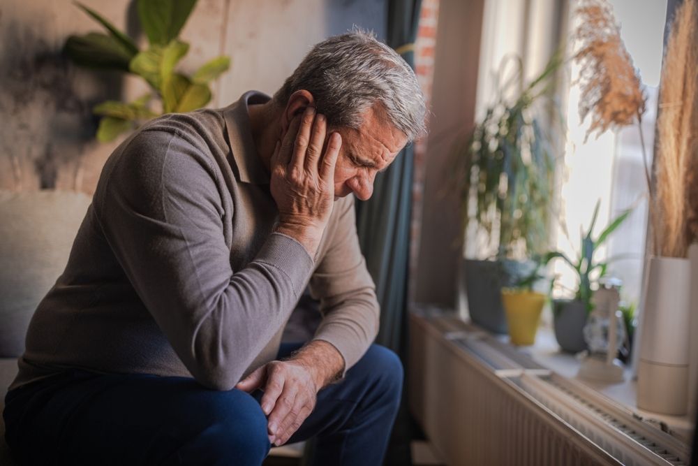 An older man sits indoors with his head resting on his hand, looking down with a distressed expression, symbolizing depression and emotional struggle.