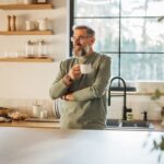 Smiling middle-aged man standing in a bright kitchen holding a coffee cup, appearing relaxed and content, symbolizing calm, routine, and mental well-being.