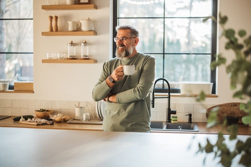 Smiling middle-aged man standing in a bright kitchen holding a coffee cup, appearing relaxed and content, symbolizing calm, routine, and mental well-being.