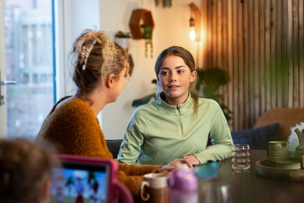 Teen girl talking with an adult at a table in a cozy home setting, appearing engaged in a serious conversation