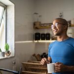 Smiling man standing in a kitchen holding a mug, looking out an open window with sunlight streaming in and small plants on the sill.