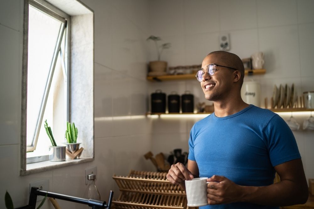 Smiling man standing in a kitchen holding a mug, looking out an open window with sunlight streaming in and small plants on the sill.