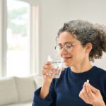 A woman with curly hair and glasses smiles as she prepares to take a pill with a glass of water in a bright, comfortable living room.