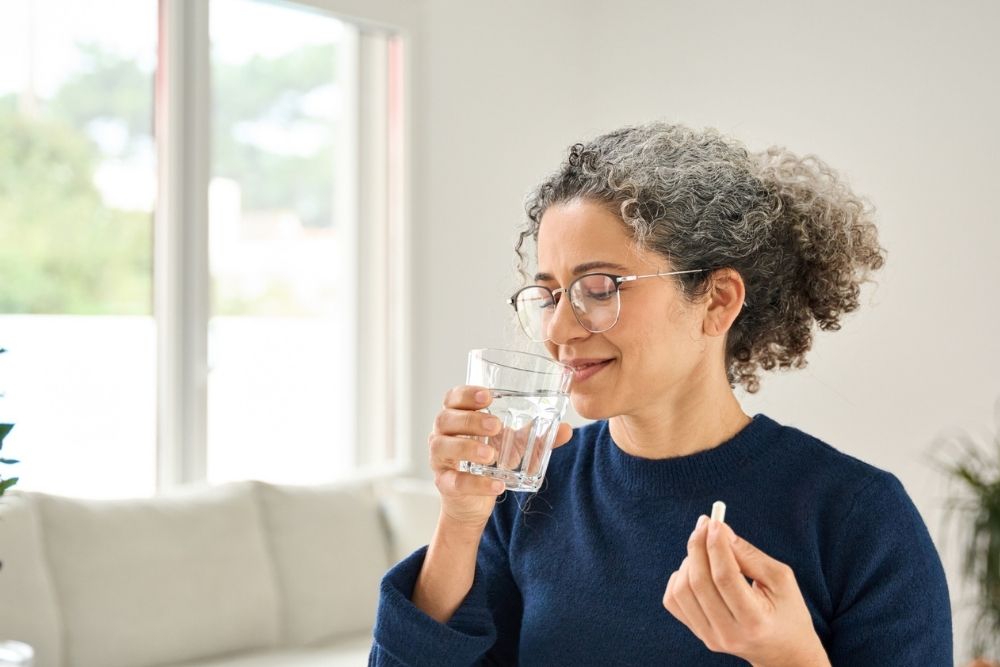 A woman with curly hair and glasses smiles as she prepares to take a pill with a glass of water in a bright, comfortable living room.