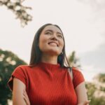 A young woman in a red shirt smiles while standing outdoors, with trees and sky in the background.