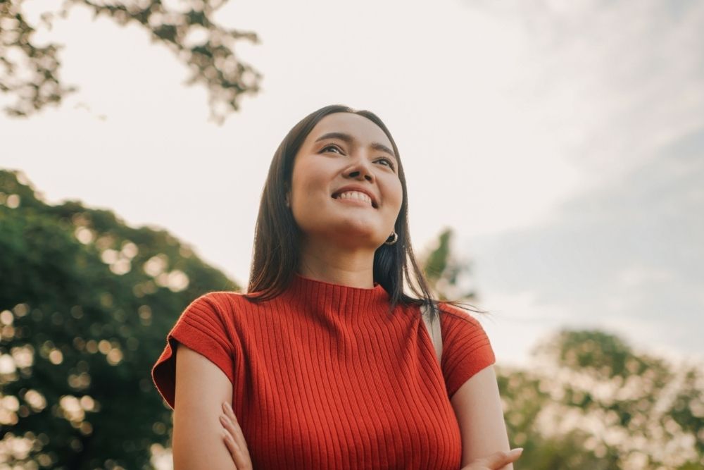 A young woman in a red shirt smiles while standing outdoors, with trees and sky in the background.