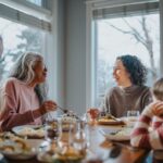 A multigenerational family sits together at a dining table during the holidays, smiling and enjoying a meal in a warmly lit home.