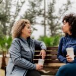 Two women sitting on a bench outdoors, holding coffee cups and having a serious conversation.