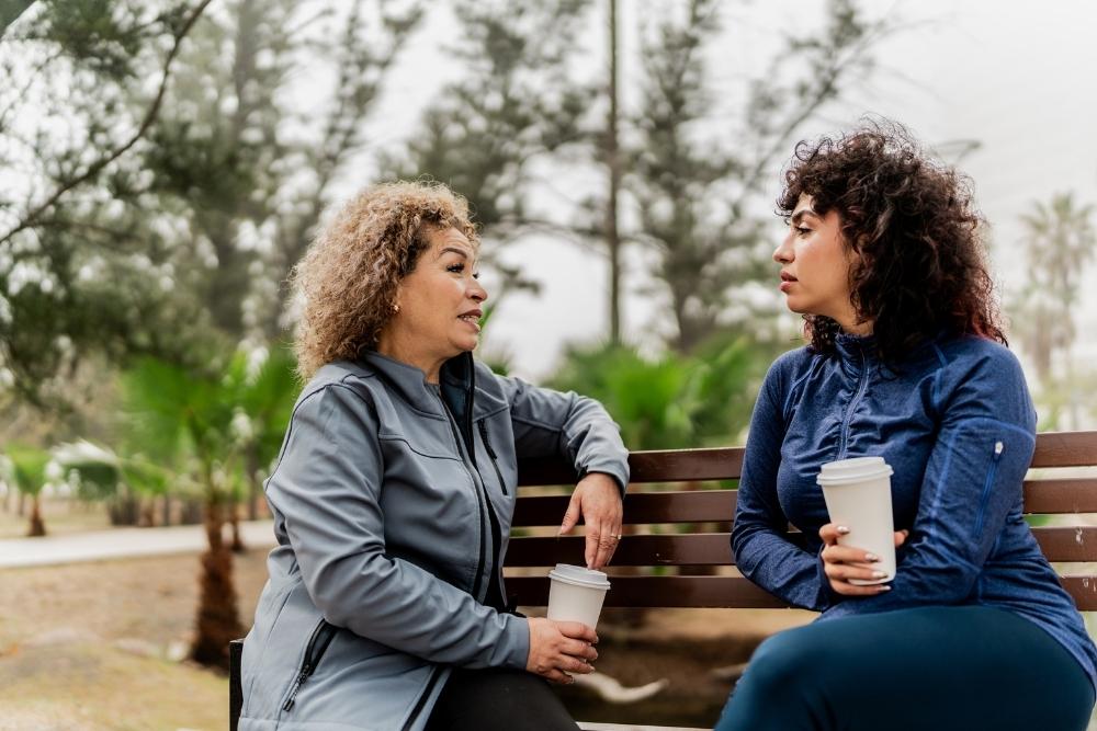 Two women sitting on a bench outdoors, holding coffee cups and having a serious conversation.
