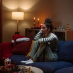 A woman sits alone on a couch in a dimly lit living room decorated for the holidays, looking thoughtful and sad as candles and festive decorations glow softly around her.