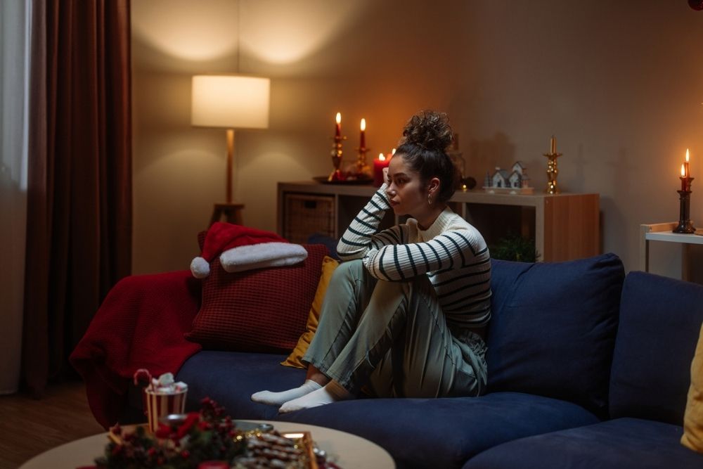A woman sits alone on a couch in a dimly lit living room decorated for the holidays, looking thoughtful and sad as candles and festive decorations glow softly around her.