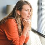 A woman rests her chin on her hands while gazing out a window, appearing deep in thought or emotionally overwhelmed in a quiet room.