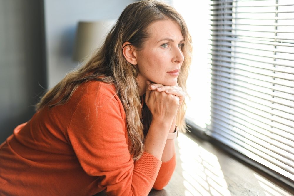 A woman rests her chin on her hands while gazing out a window, appearing deep in thought or emotionally overwhelmed in a quiet room.