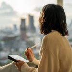 Person sitting by a window with a notebook and pen, reflecting quietly while looking out at a city skyline.