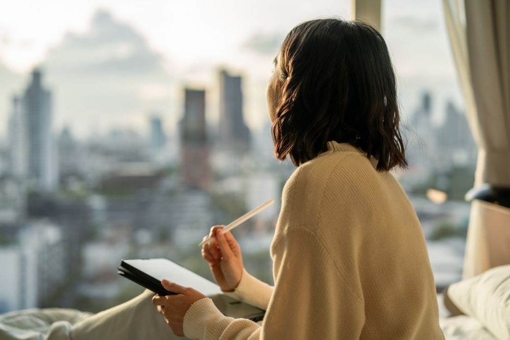 Person sitting by a window with a notebook and pen, reflecting quietly while looking out at a city skyline.