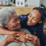 Peak Behavioral - How to Create a Supportive Home Environment for Mental Wellness - Edited. A yong woman hugs an older woman on a couch. They both look at each other smiling.