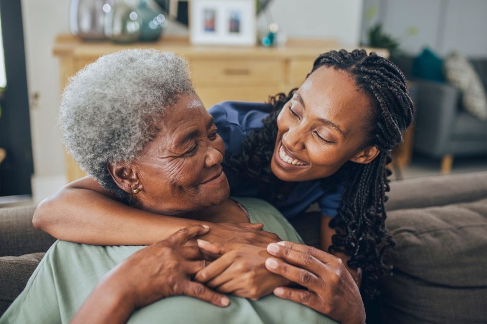 Peak Behavioral - How to Create a Supportive Home Environment for Mental Wellness - Edited. A yong woman hugs an older woman on a couch. They both look at each other smiling.